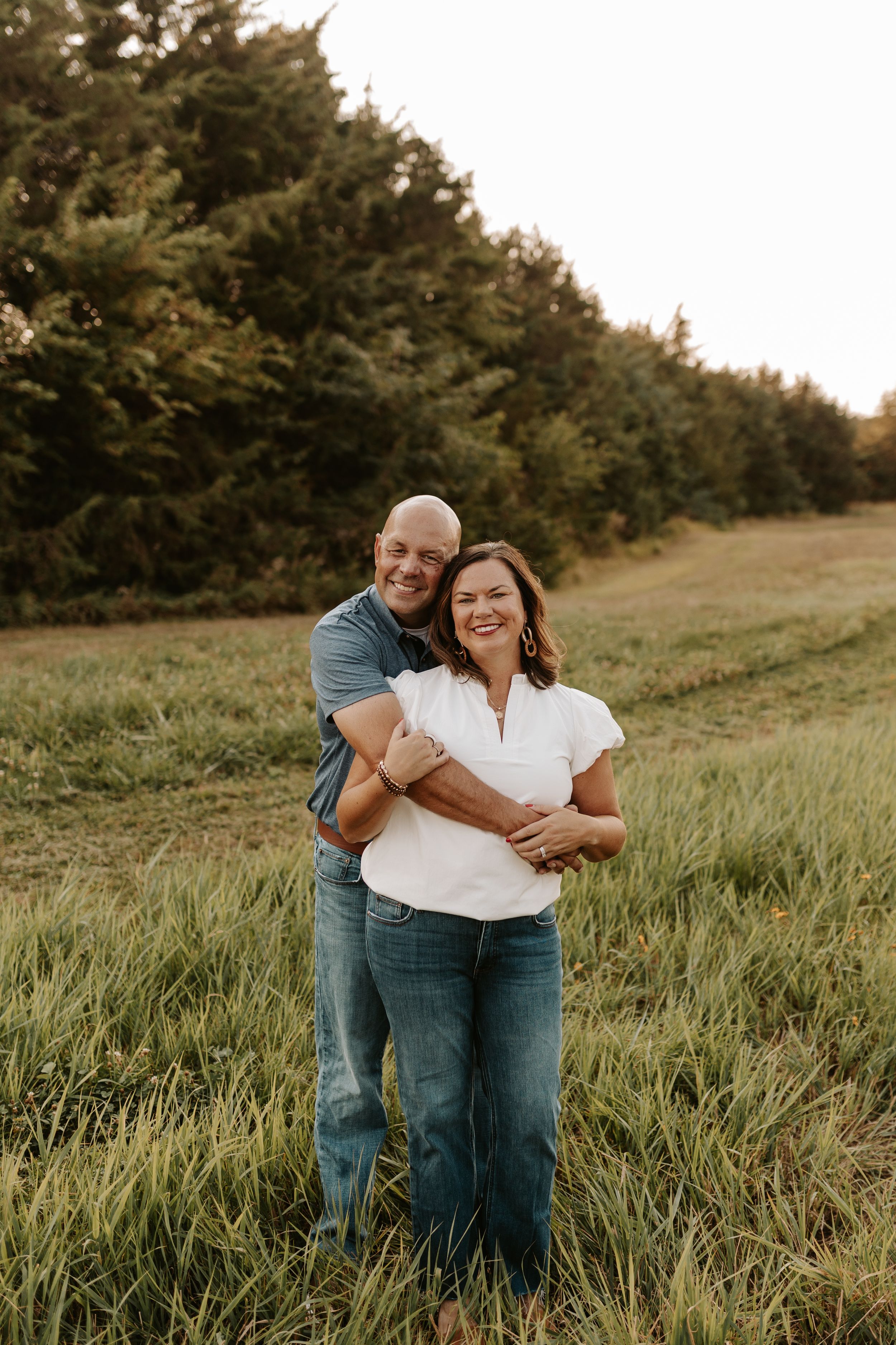 A smiling couple embraces in a lush green field with trees in the background during golden hour.