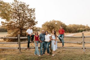 Family of six posing by a rustic wooden fence in a golden field during sunset, wearing coordinated casual outfits.