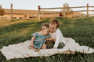 Two young siblings sit on a blanket in a sunny green field, with an older boy affectionately leaning toward a baby girl.