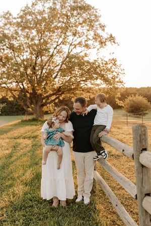 Family of four posing by a wooden fence in a golden field at sunset, parents holding young children near a large oak tree.