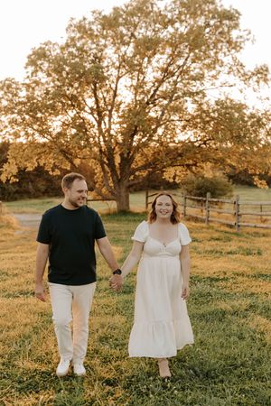 Couple holding hands in a golden hour field, man in black tee, woman in white dress, large oak tree behind them.