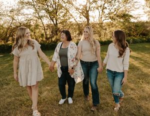 Four women holding hands walking together outdoors in golden hour light, wearing casual outfits in a green park setting.