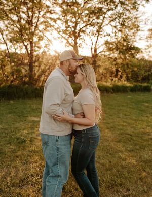 Couple sharing a romantic moment at golden hour in a grassy field, woman in white top and jeans, man in cap and light shirt.