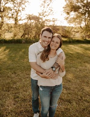 A smiling couple embraces outdoors in a sunny field, wearing casual jeans and light tops, surrounded by green trees.