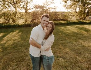 Happy couple embracing outdoors at golden hour, smiling in a lush green field with trees in the background.