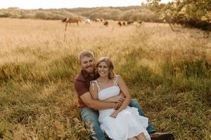 Couple sitting together in golden field at sunset, woman in white dress leaning on man in red shirt, cows grazing behind them.