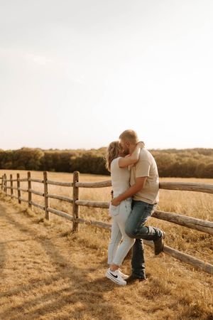Couple embracing and kissing by a rustic wooden fence in a golden field at sunset.