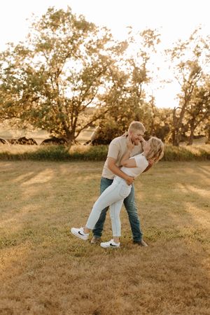 Couple sharing a romantic dip kiss in a golden hour field with trees in the background.