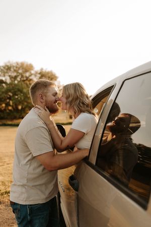 Couple sharing a romantic moment leaning in for a kiss beside a silver car outdoors at golden hour.