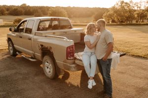Couple sharing a romantic moment leaning against a silver pickup truck tailgate at golden hour on a rural farm.