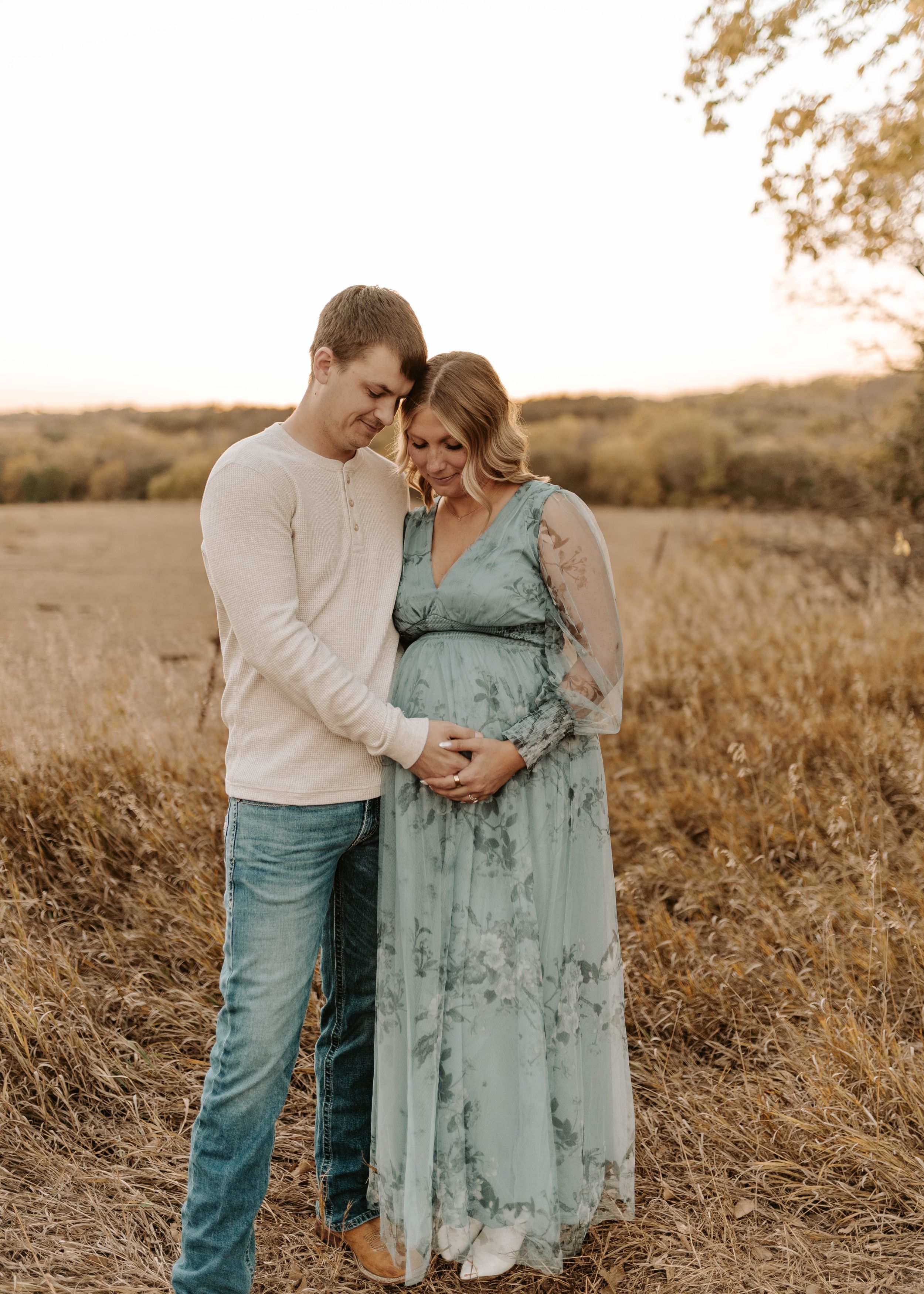 Couple sharing a tender moment in a golden field, woman in floral maternity dress cradling her baby bump at sunset.