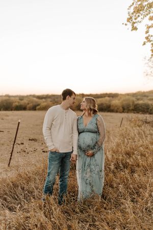 Couple sharing a tender moment in a golden field at sunset, woman in floral maternity dress cradling baby bump.