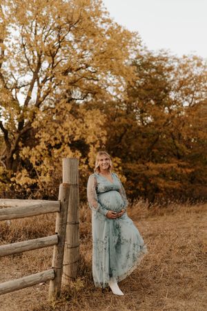 Pregnant woman in teal floral dress leaning on wooden fence surrounded by golden autumn trees.