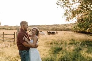 Couple embracing in golden hour sunlight in a rustic farm field with cattle and wooden fence in background.