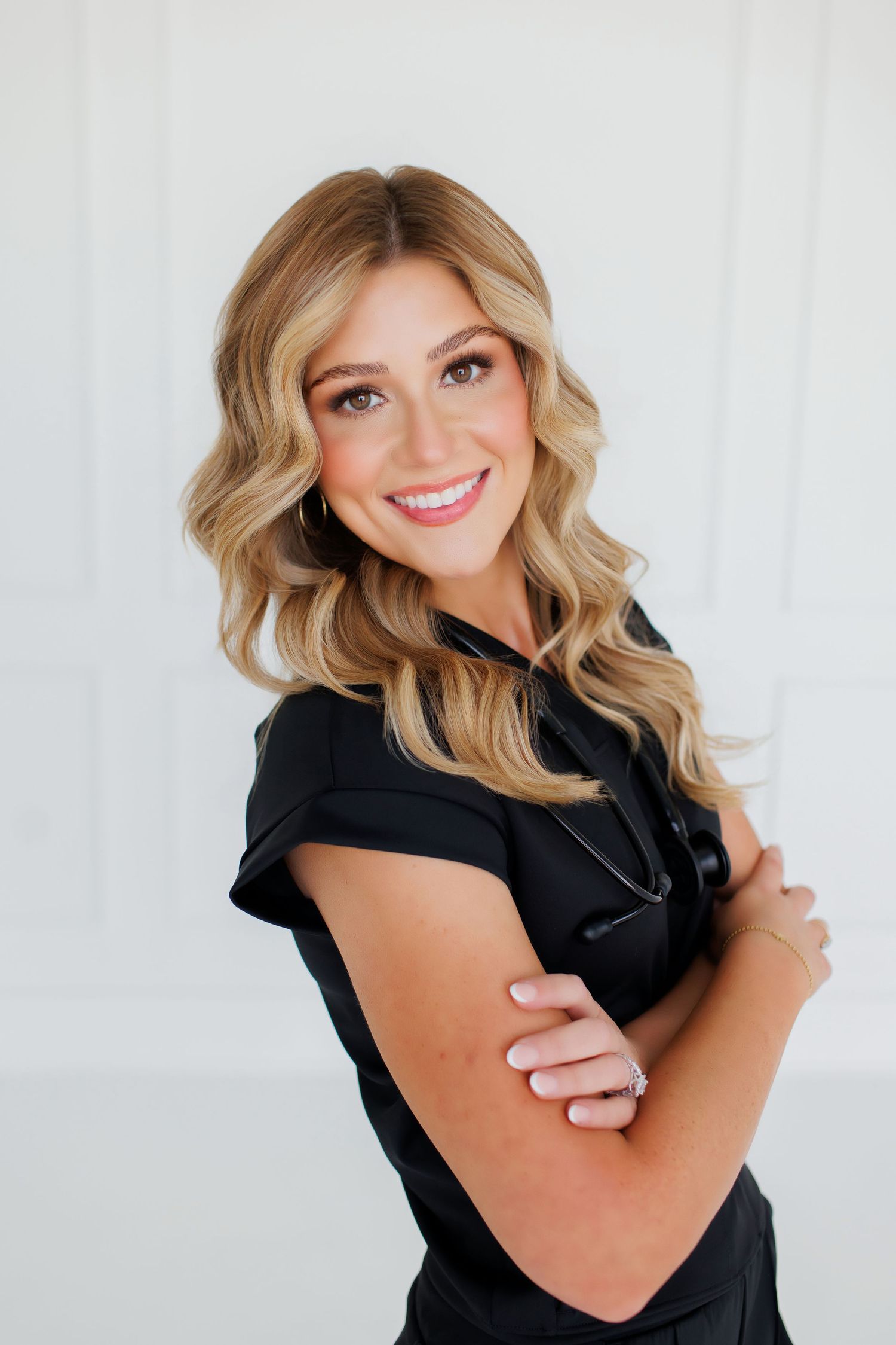 Smiling blonde woman in white blazer sitting on sofa, wearing gold watch and ring, with wavy hair and elegant style.