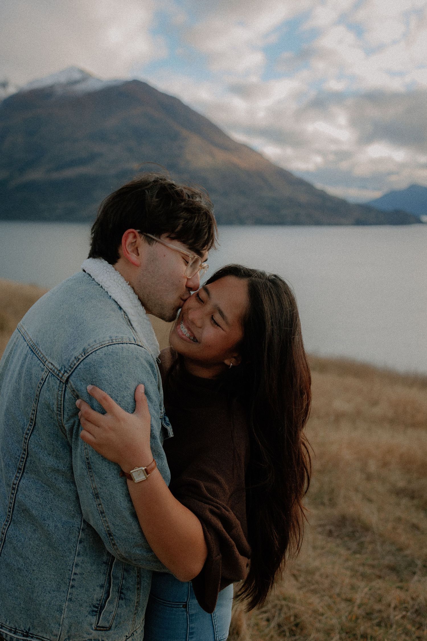 A couple embraces and shares a tender kiss outdoors near a scenic lake with mountains and cloudy skies.