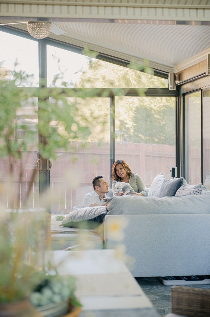 Couple relaxing together on a large white sofa in a bright sunroom with floor-to-ceiling windows overlooking a garden.