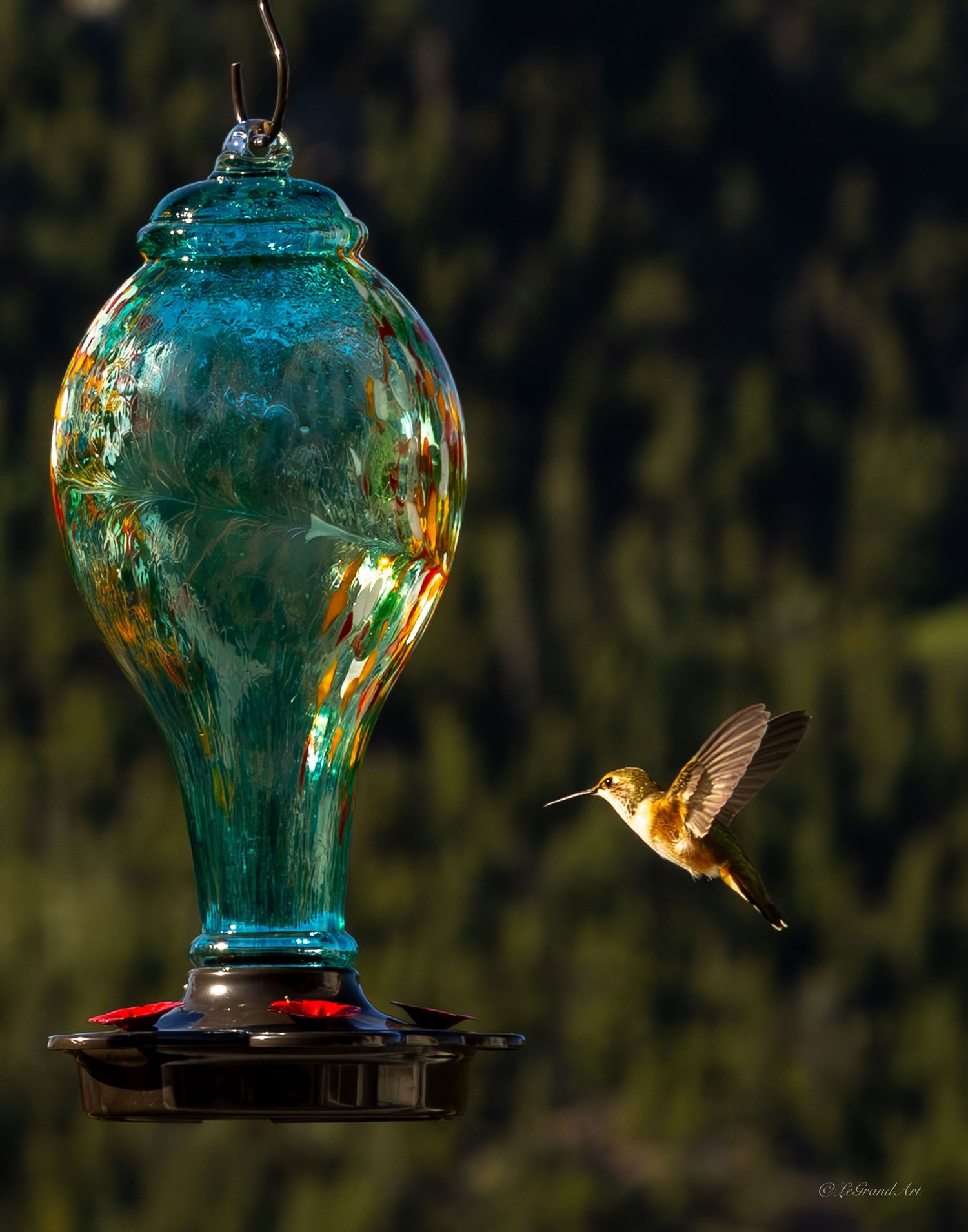A hummingbird hovering near a decorative teal glass hummingbird feeder.