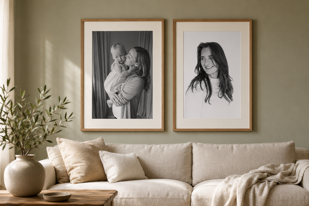 Black and white framed family portrait and woman portrait hanging on sage green wall above beige linen sofa with cushions.
