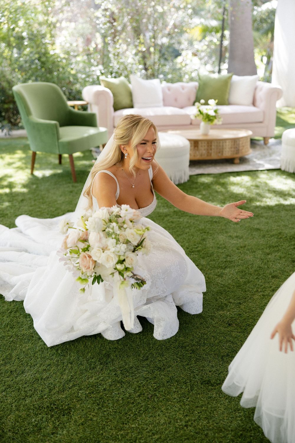 Smiling bride in white gown kneeling on grass holding bouquet of white and pink flowers at outdoor wedding reception.