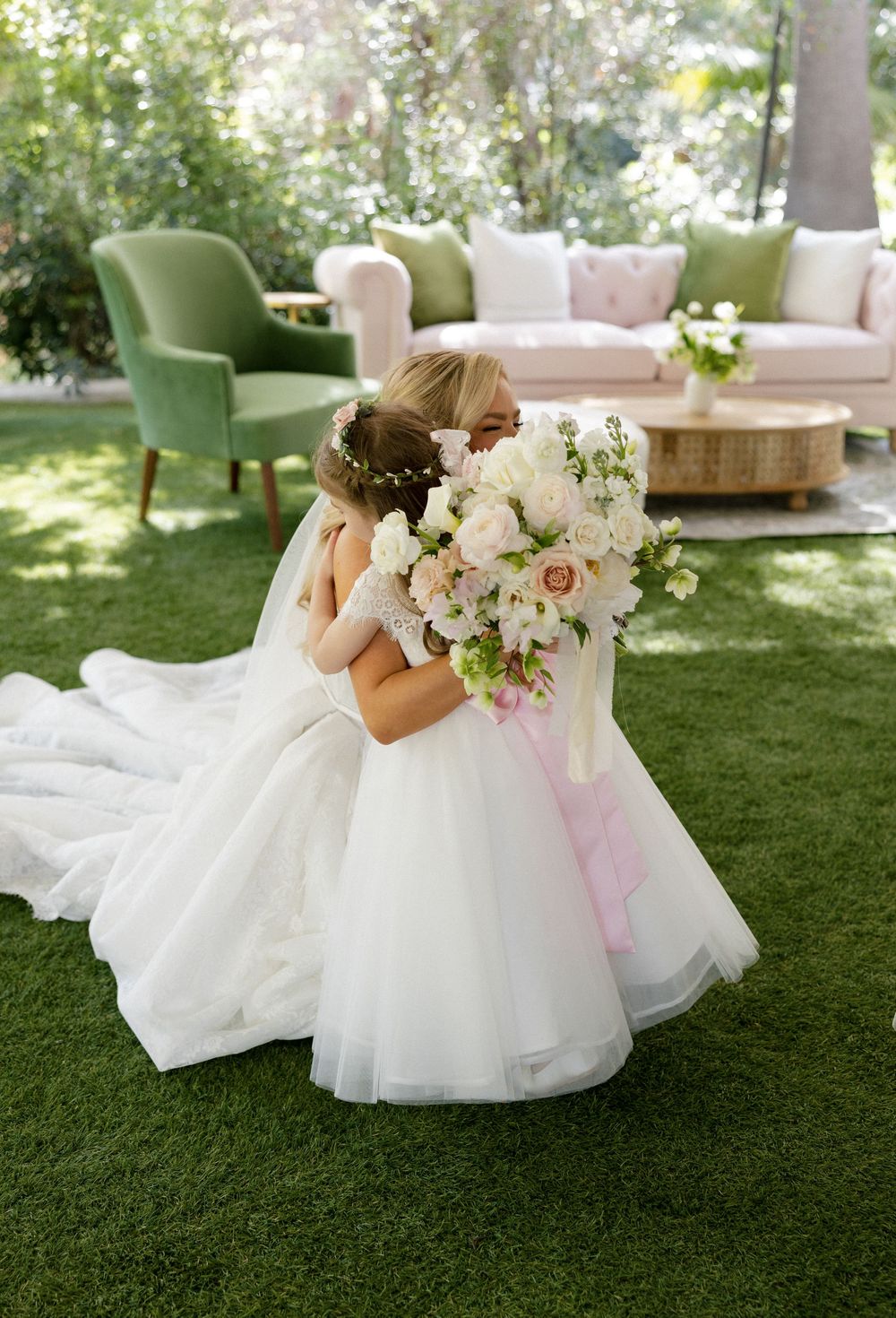 Bride in white gown kneeling to hug flower girl holding bouquet on green lawn at outdoor wedding.