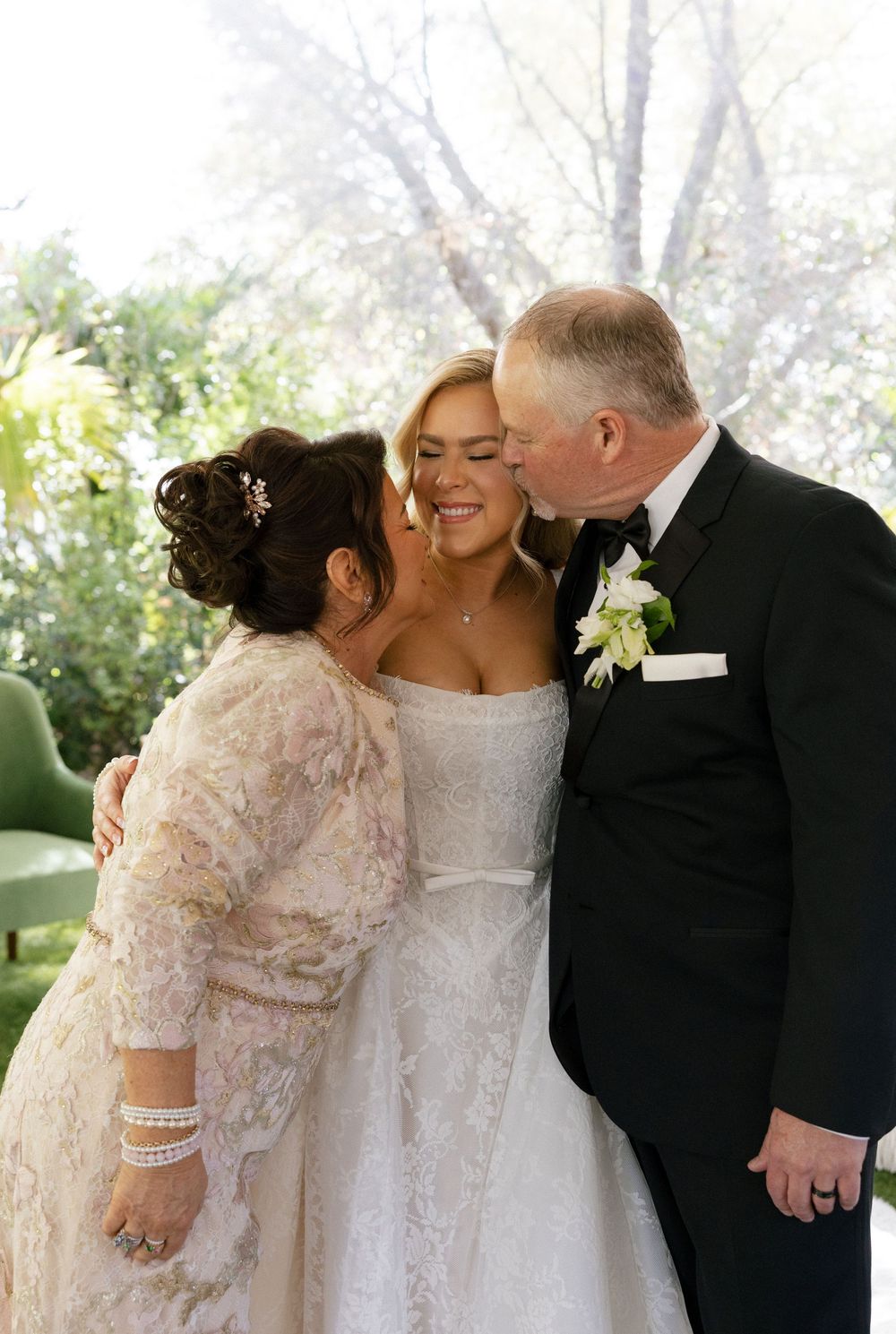 Bride in white lace gown receives kisses from parents on wedding day outdoors.