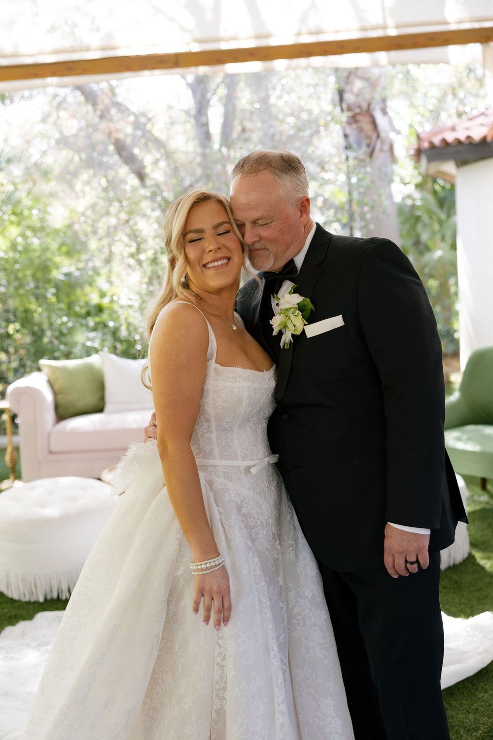 A smiling bride in a white lace gown poses with a man in a black tuxedo at an outdoor wedding ceremony.