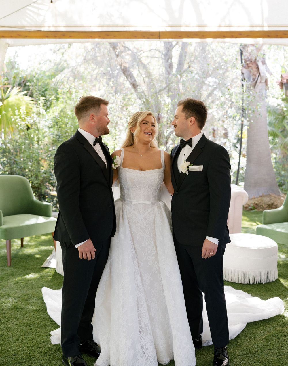 Bride in white gown stands with two grooms in tuxedos at outdoor wedding ceremony on green lawn.
