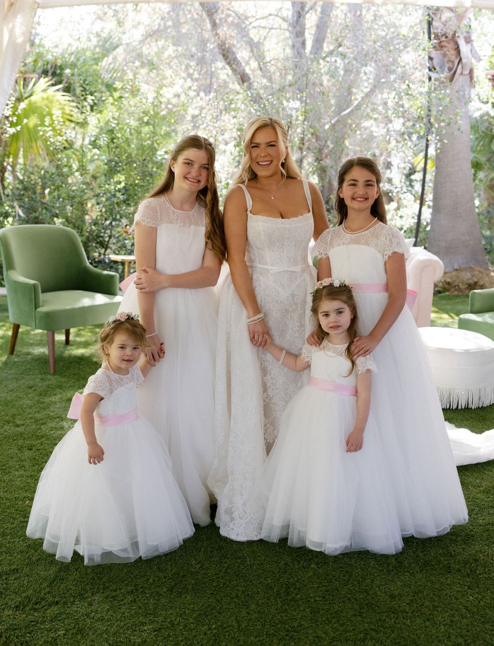 Bride in lace wedding gown poses with four flower girls in white dresses with pink sashes outdoors.