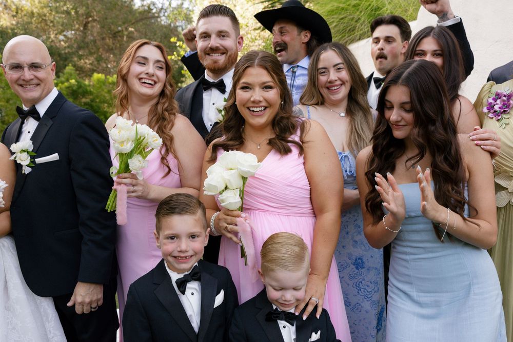 Wedding party group photo with bridesmaids in pink dresses, groomsmen in suits, and children in formal attire outdoors.