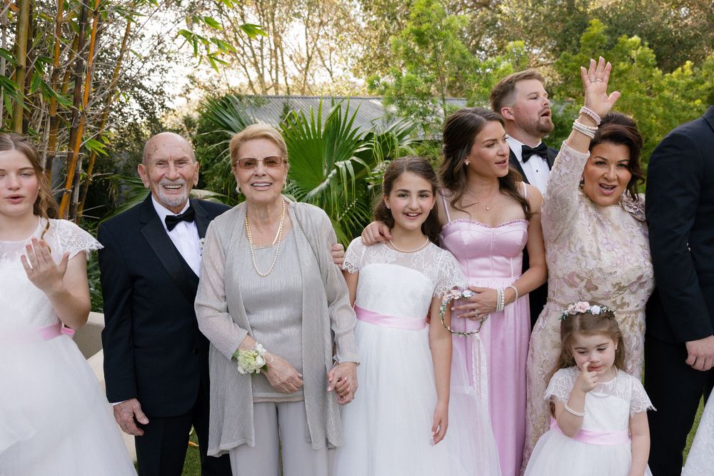 Family group photo at a wedding with guests in formal attire smiling and waving outdoors.