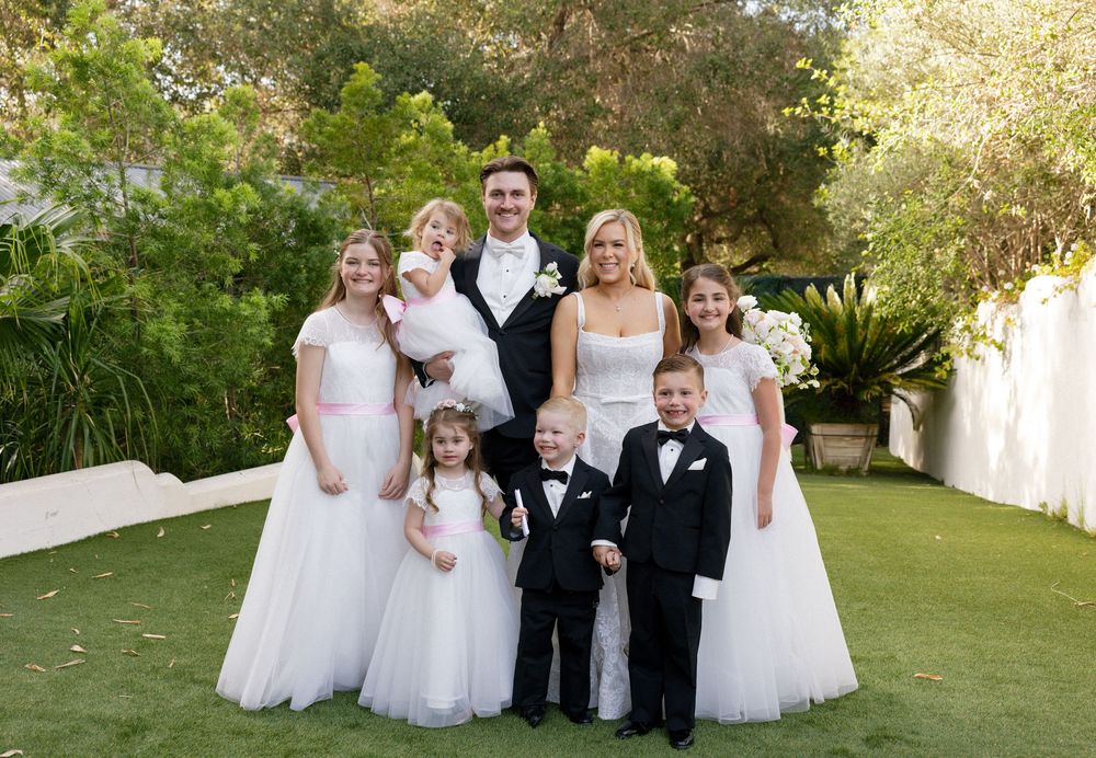 Wedding party group photo with bride, groom, flower girls, and ring bearer posing outdoors on green lawn.