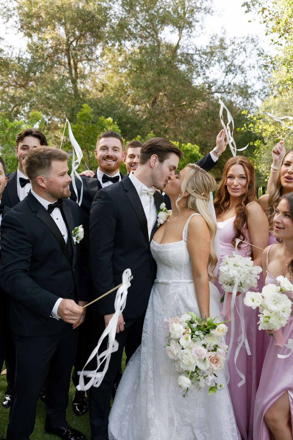 Bride and groom share a kiss surrounded by smiling wedding party in black tuxedos and pink bridesmaid dresses outdoors.