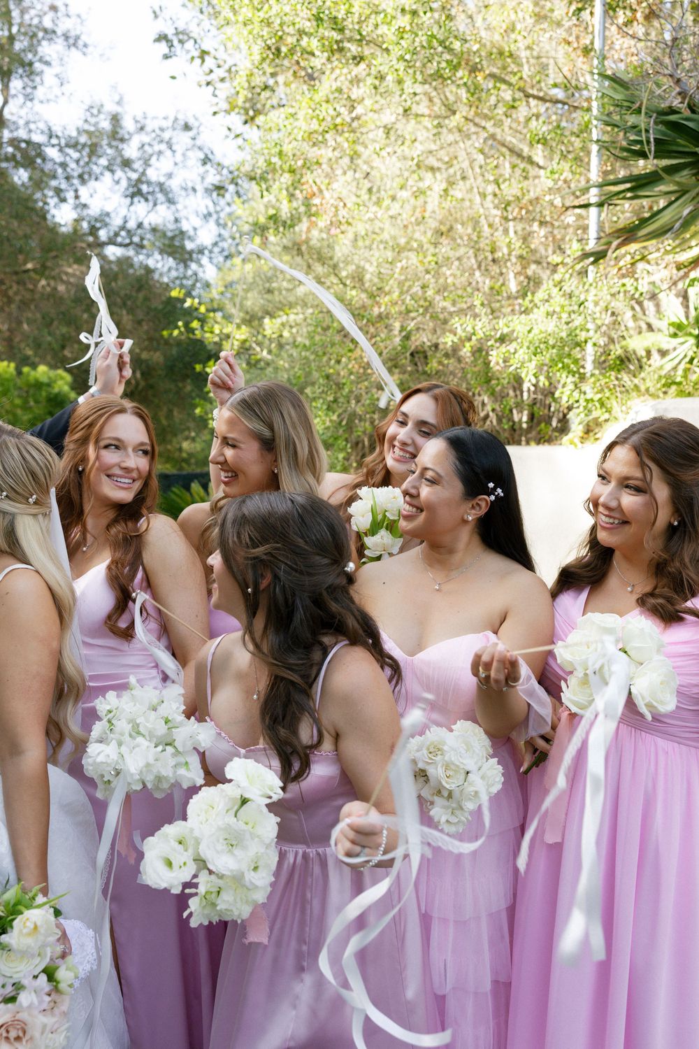 Bridesmaids in pink dresses laugh and celebrate together holding white floral bouquets and ribbon wands outdoors.
