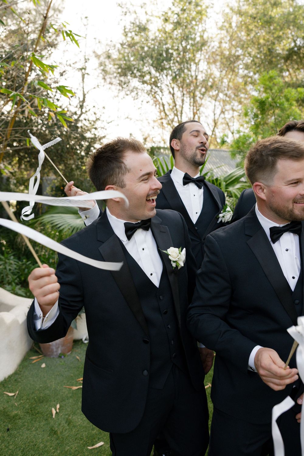 Groomsmen in black tuxedos celebrate outdoors waving white ribbon wands, laughing joyfully at a wedding reception.
