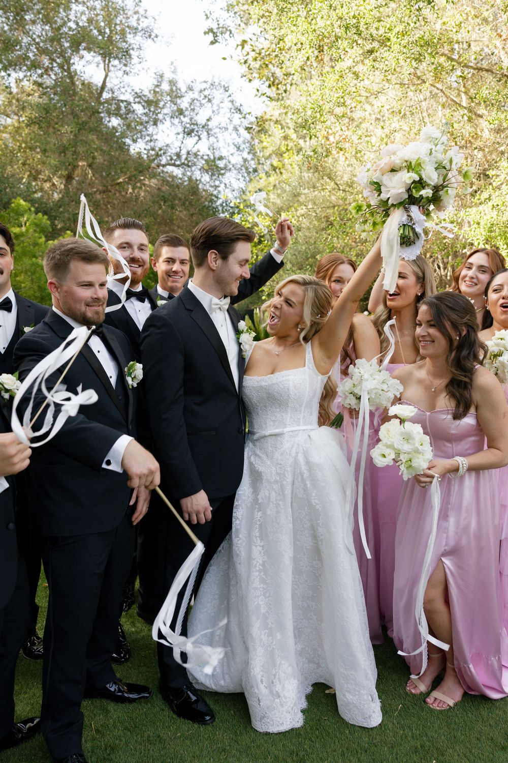 Joyful wedding party celebrating outdoors with bride in white gown holding bouquet aloft, bridesmaids in pink, groomsmen in black tuxedos.
