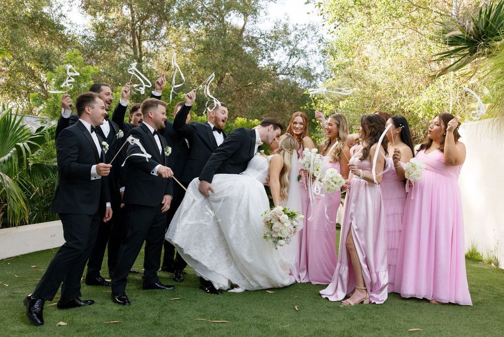 Wedding party celebrates outdoors with bride and groom kissing, groomsmen in tuxedos and bridesmaids in pink gowns.