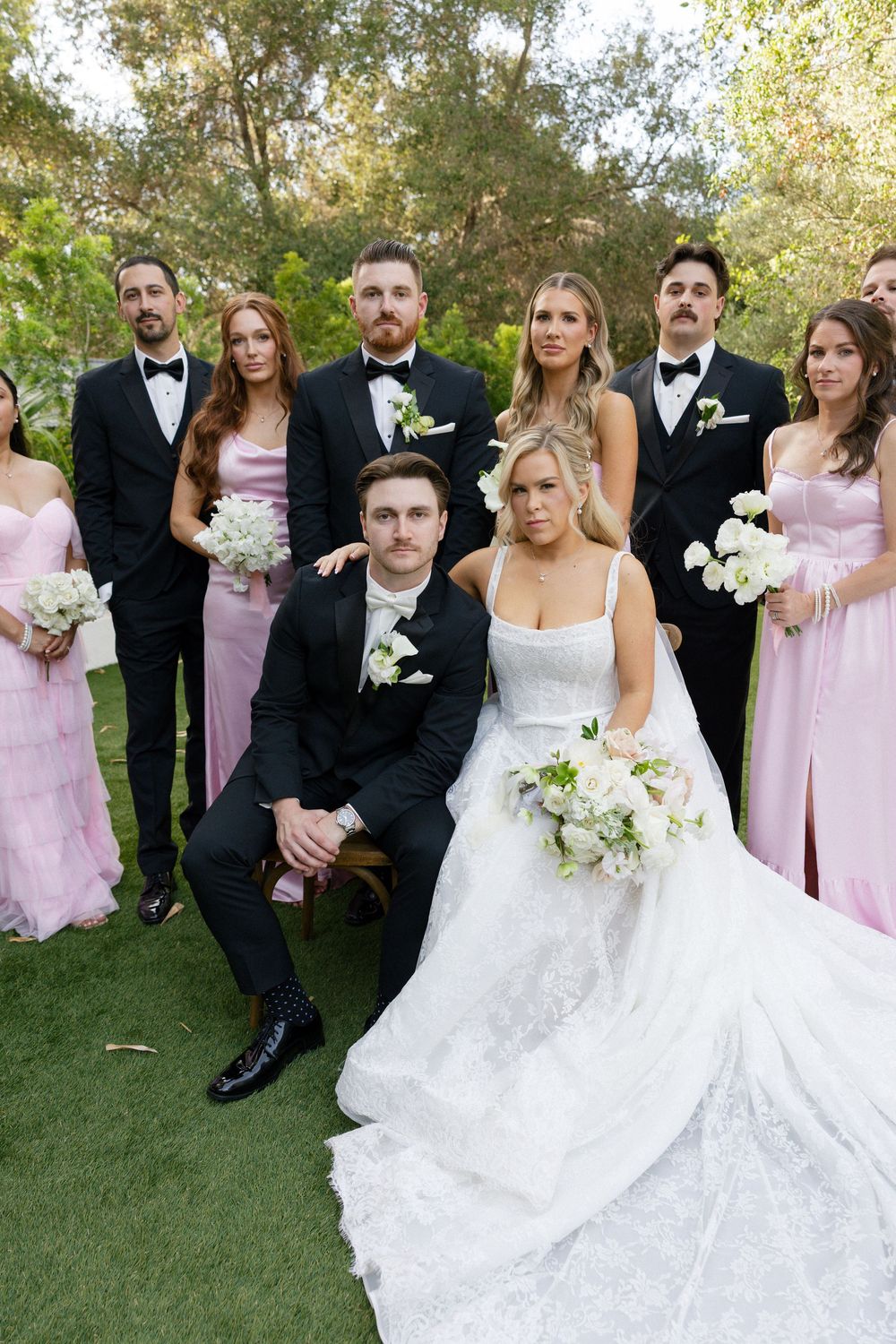 Elegant wedding party portrait outdoors with bride and groom seated, surrounded by bridesmaids in pink gowns and groomsmen in black tuxedos.