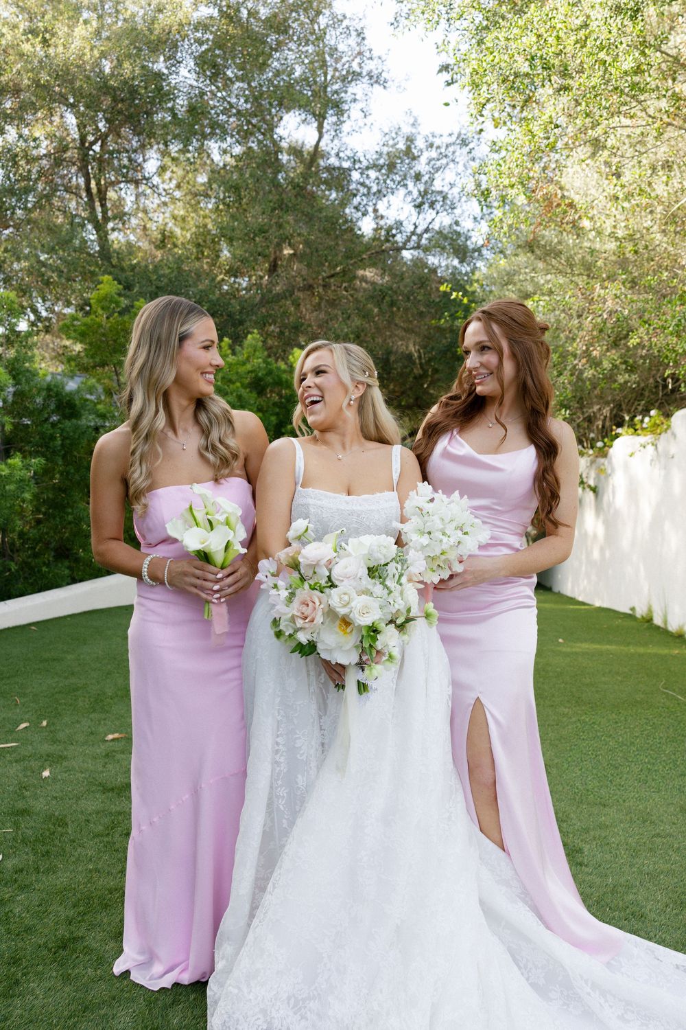 Bride in white lace gown laughs joyfully with two bridesmaids in pink satin dresses holding white bouquets outdoors.
