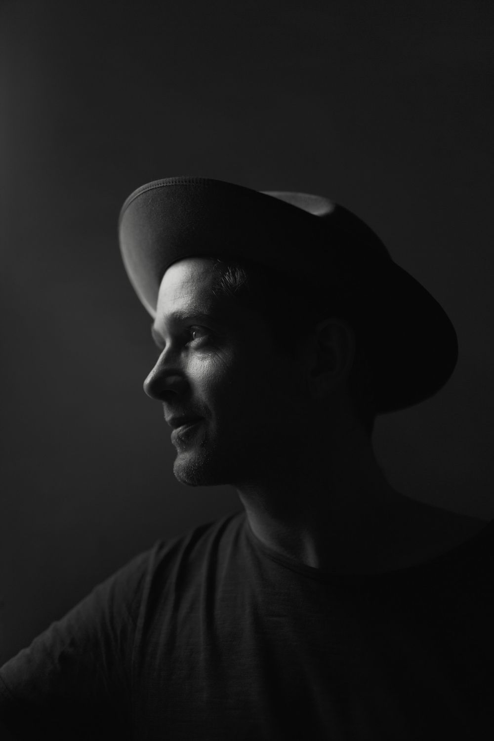 Moody black and white profile portrait of figure in hat against dark background creating dramatic lighting.
