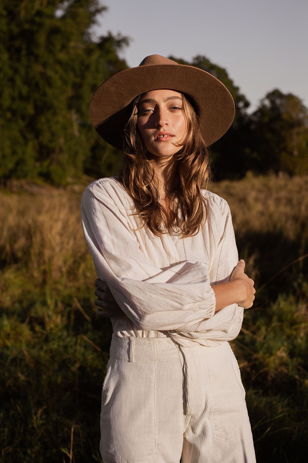 Woman in white linen dress and wide-brim hat standing in golden hour sunlight in a natural field setting.