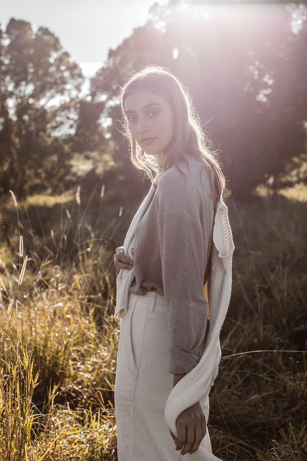 Woman in beige cardigan and trousers standing in sunlit field with trees, golden backlight creating warm glow.