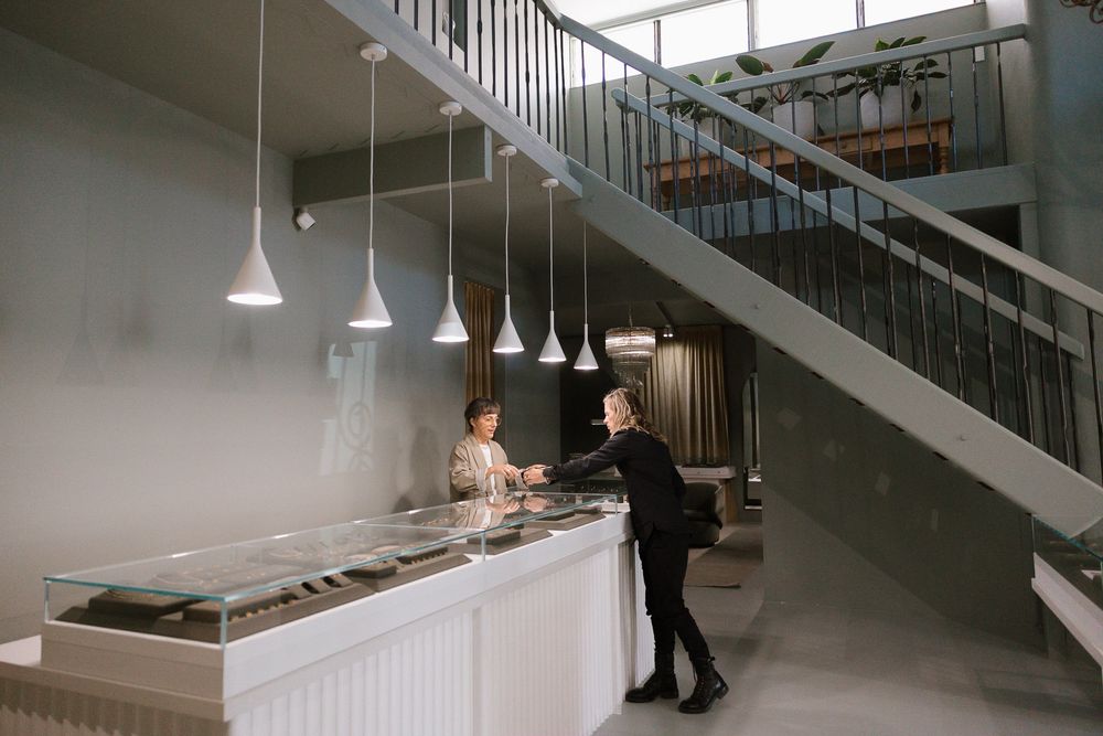 Two people at a modern jewelry store counter with pendant lights and an elegant staircase in the background.