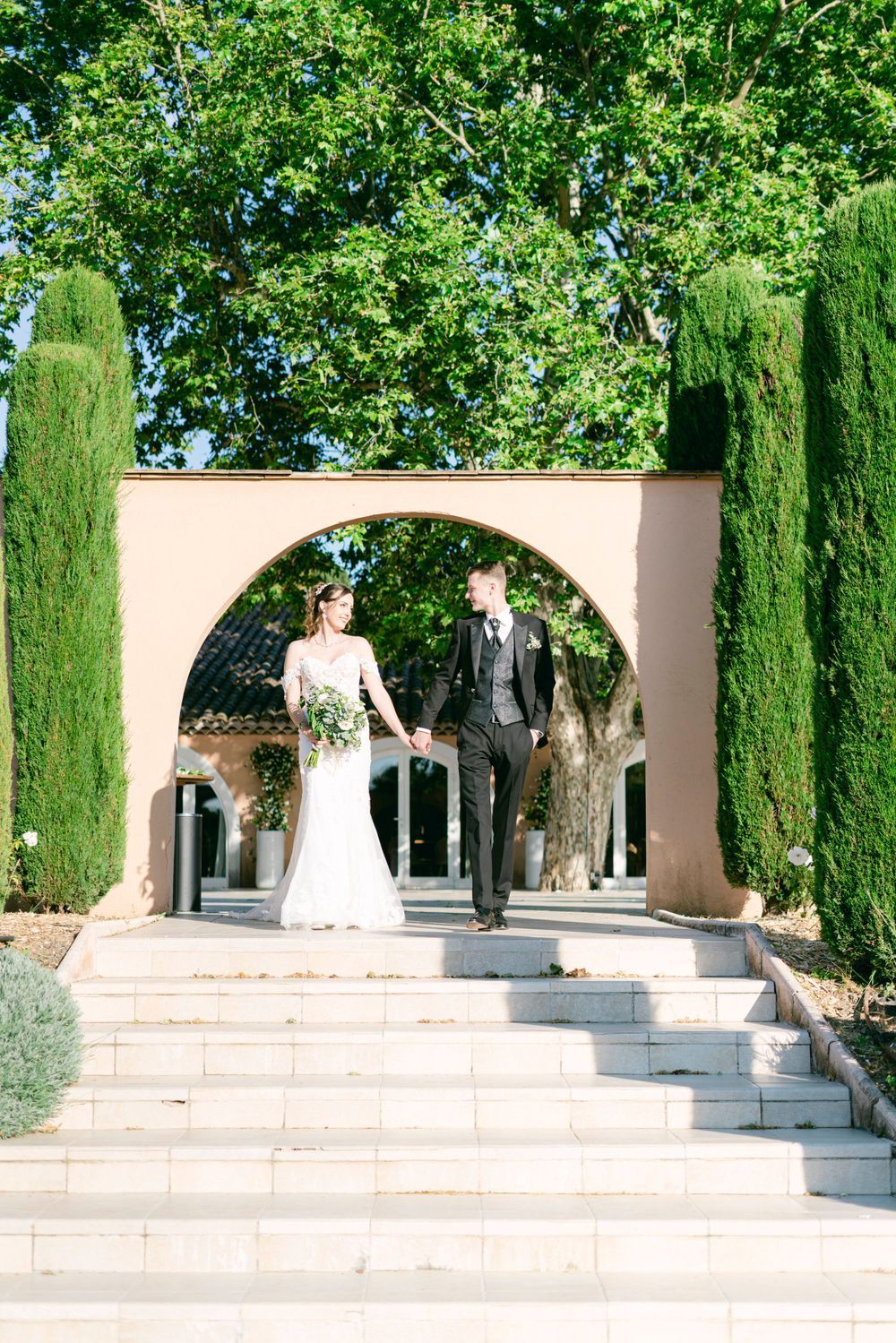 Bride and groom holding hands walking down steps through a circular arch surrounded by cypress trees.