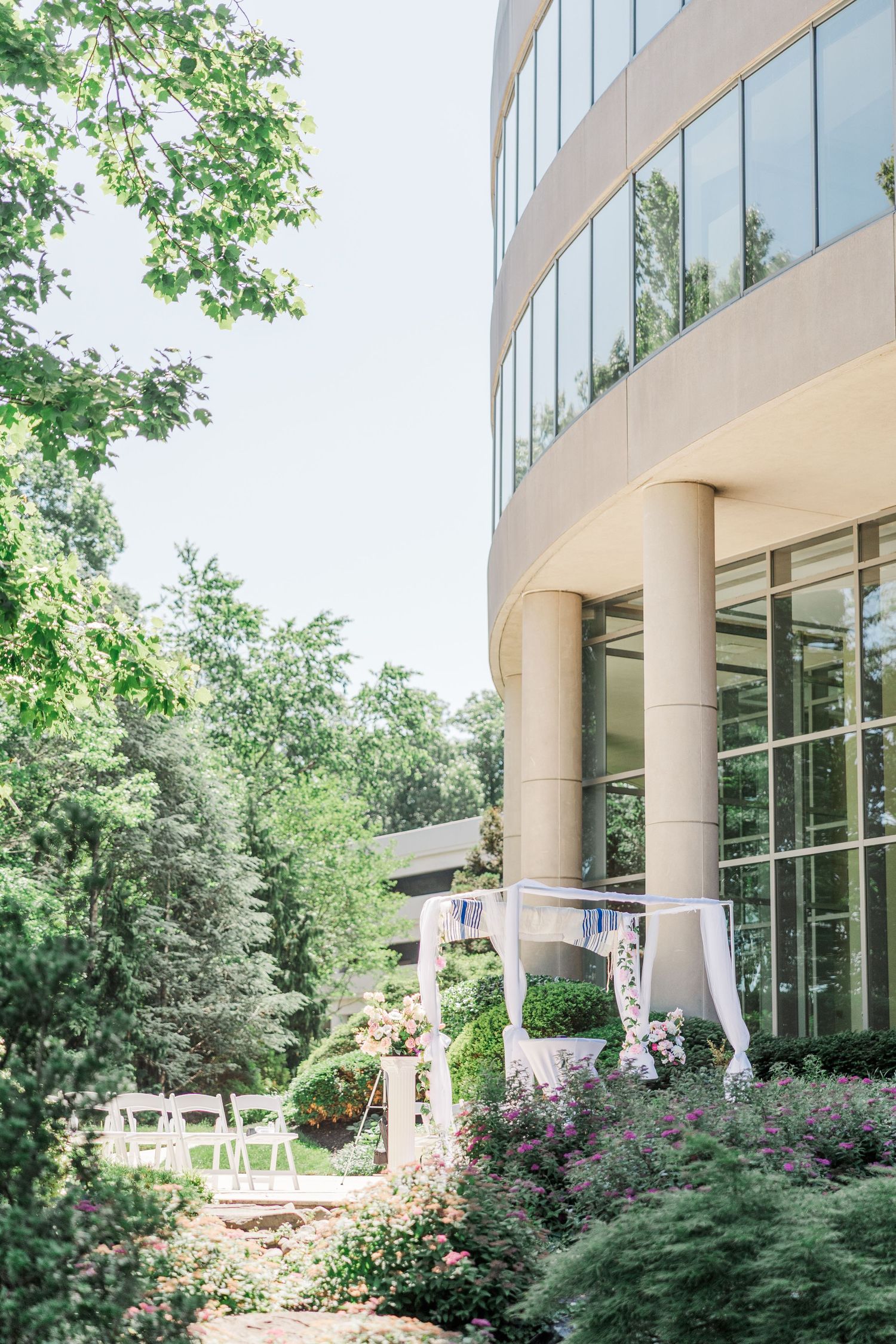 Outdoor wedding ceremony setup with white chairs and floral arch beside a modern glass building surrounded by lush greenery.