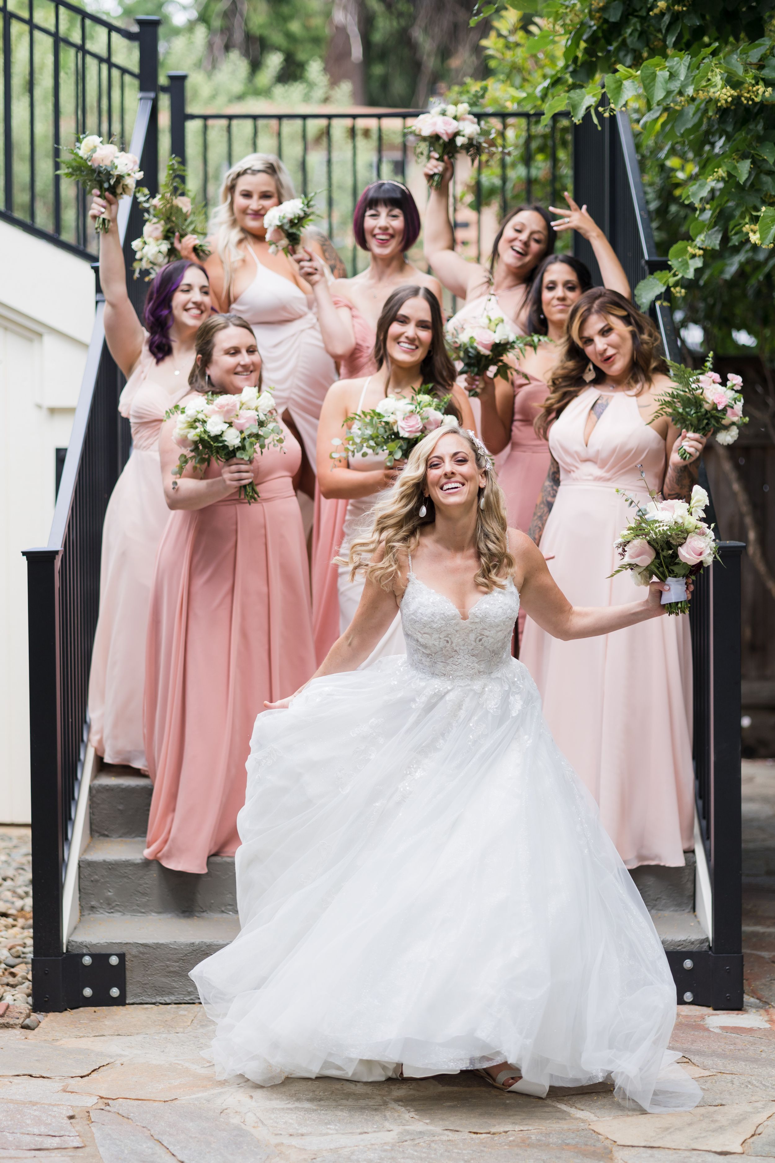 Bride in white gown poses with bridesmaids in pink dresses holding bouquets on outdoor staircase.