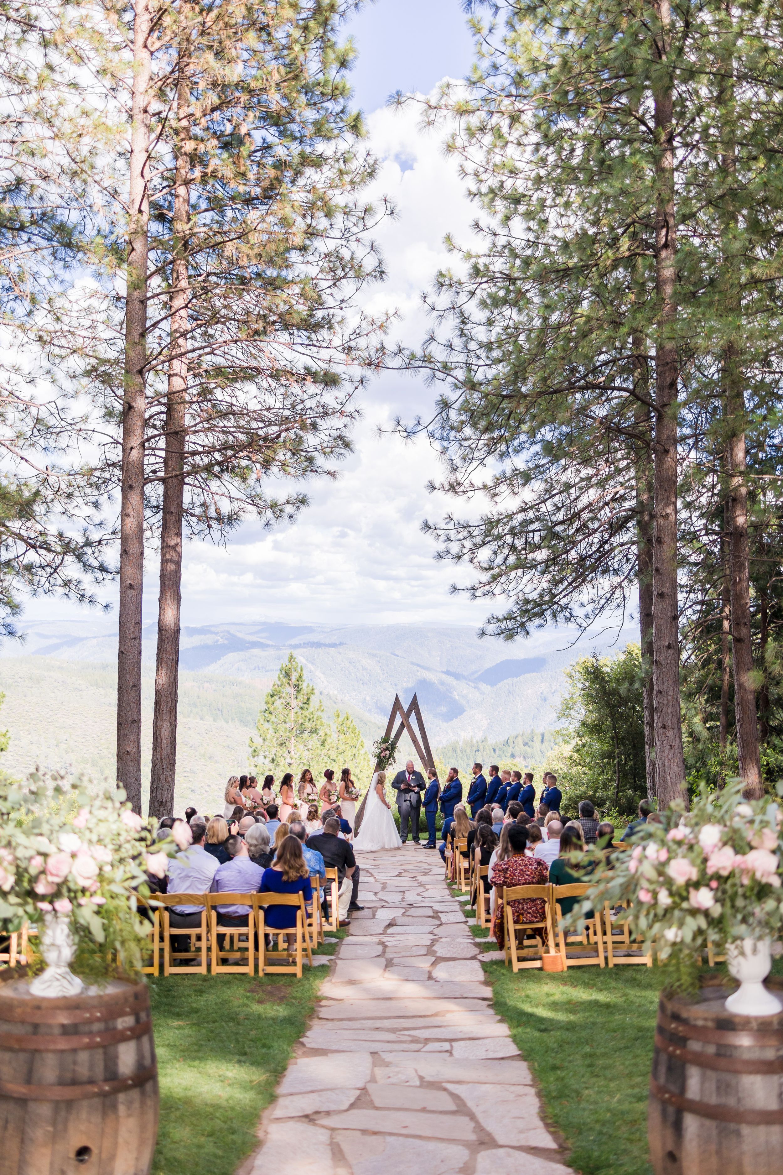 Outdoor wedding ceremony with wooden arch, stone aisle, tall pines, floral arrangements on barrels, and mountain views.