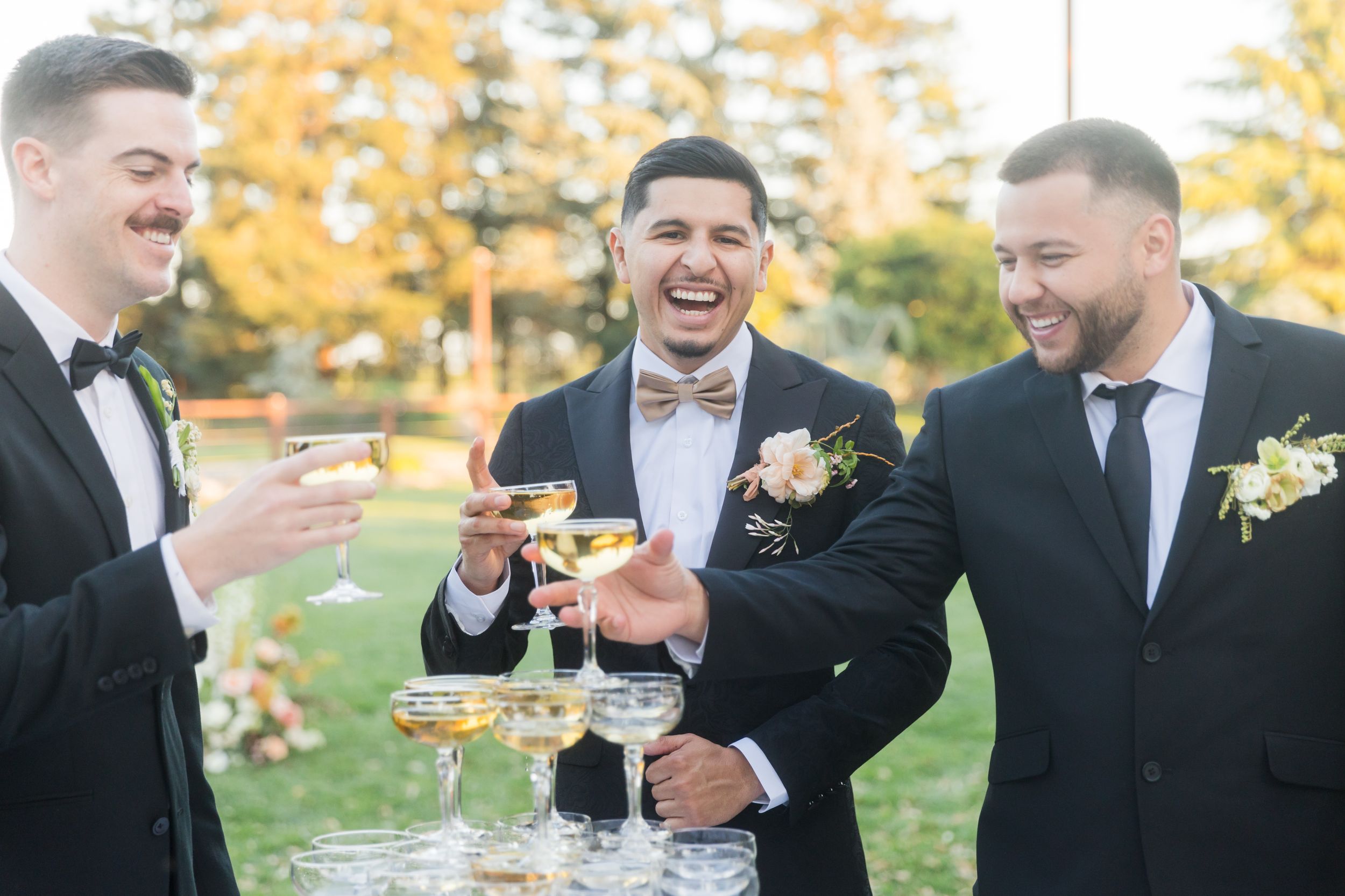 Three groomsmen in tuxedos laughing and toasting with champagne glasses at an outdoor wedding celebration.