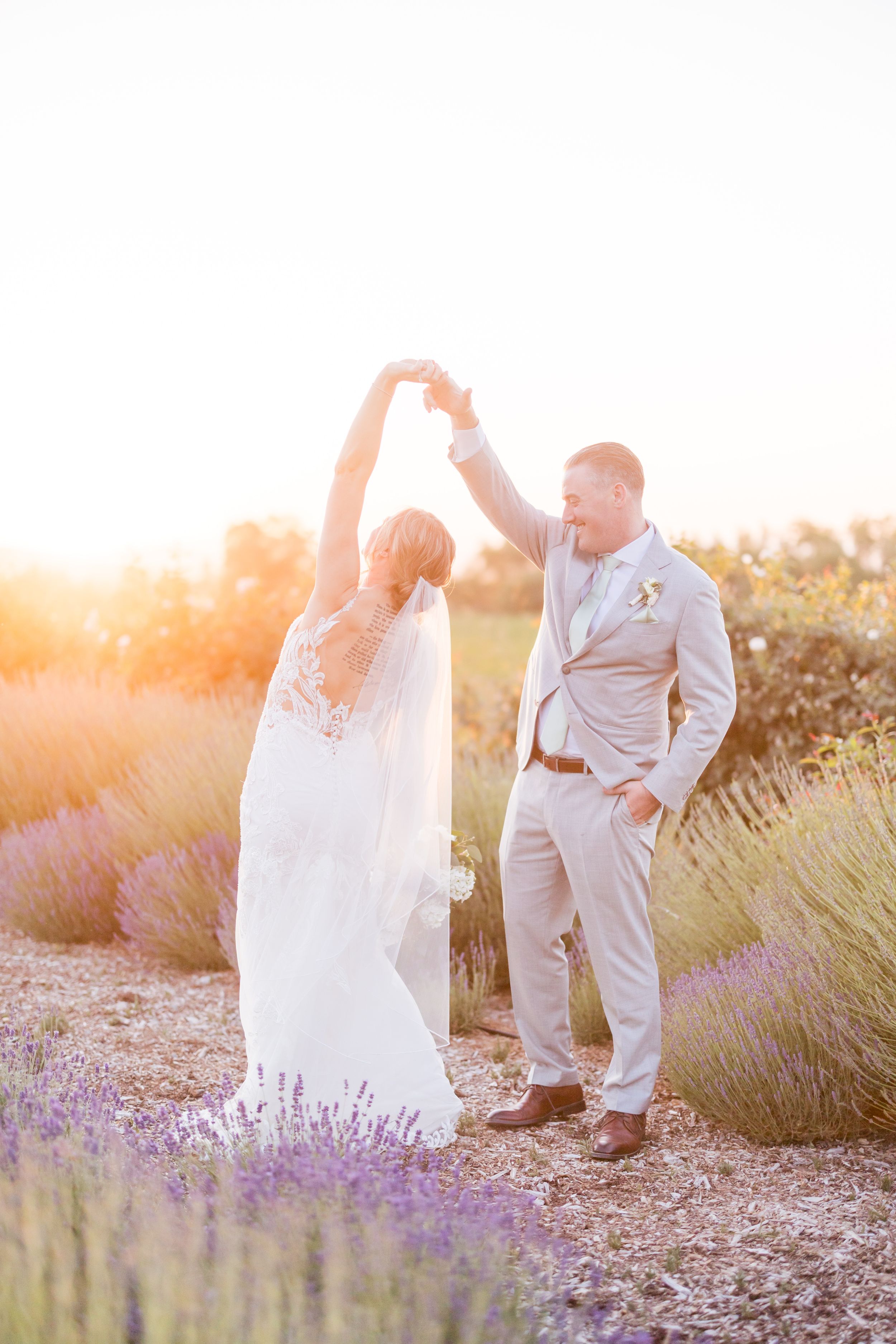 Bride and groom dancing in a lavender field at golden hour, bride in lace gown with veil, groom in gray suit.
