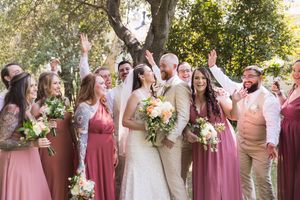 Bride and groom surrounded by bridesmaids in dusty rose gowns and groomsmen celebrating outdoors under trees.